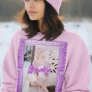 May include: A pink sweatshirt with a purple frame featuring a photo of Marilyn Monroe. The sweatshirt has a pink beanie with a pom-pom and an anchor graphic with the text "I'd rather be sailing".