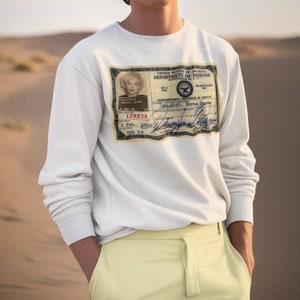 Könnte beinhalten: Ein weißer Langarm-Pullover mit einer aufgedruckten ID-Karte im Vintage-Stil auf der Vorderseite. Die ID-Karte zeigt ein Schwarzweißfoto einer Frau mit dem Text "DIMENDID NORMA JEANE" und "129278". Der Text "UNITED STATES OF AMERICA DEPARTMENT OF DEFENSE" ist oben auf der Karte aufgedruckt.