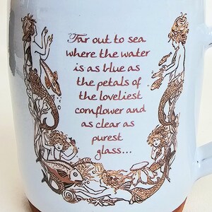 May include: White ceramic mug with a light blue glaze and a brown base. The mug features a mermaid design in brown tones, with the text "Far out to sea where the water is as blue as the petals of the loveliest cornflower and as clear as purest glass..."