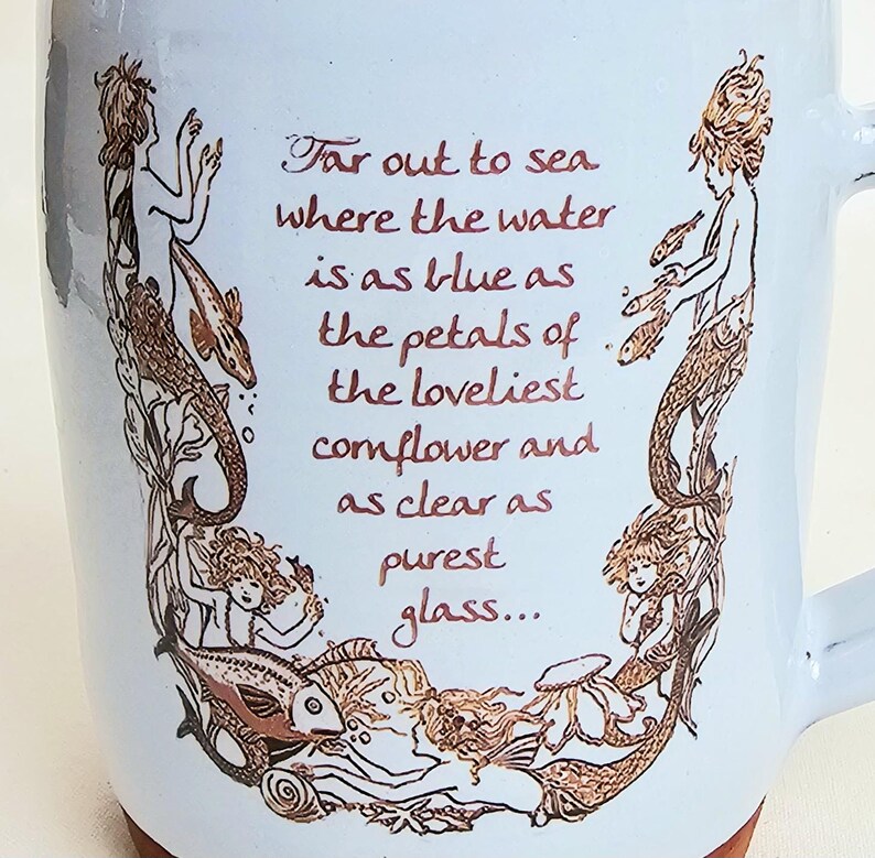 May include: White ceramic mug with a light blue glaze and a brown base. The mug features a mermaid design in brown tones, with the text "Far out to sea where the water is as blue as the petals of the loveliest cornflower and as clear as purest glass..."
