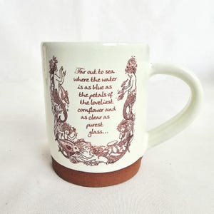 May include: Cream ceramic mug with a brown base and handle. The mug has a maroon illustration of mermaids and the text "Far out to sea where the water is as blue as the petals of the loveliest cornflower and as clear as purest glass..."