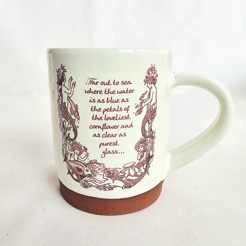 May include: Cream ceramic mug with a brown base and handle. The mug has a maroon illustration of mermaids and the text "Far out to sea where the water is as blue as the petals of the loveliest cornflower and as clear as purest glass..."