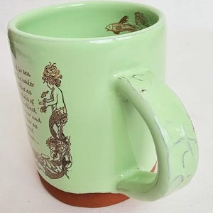 May include: A light green ceramic mug with a mermaid illustration and fish designs. The mug has a brown base and a large handle. The mermaid illustration is in brown and depicts a mermaid holding a fish. The mug has text on the side.