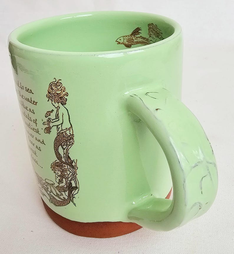 May include: A light green ceramic mug with a mermaid illustration and fish designs. The mug has a brown base and a large handle. The mermaid illustration is in brown and depicts a mermaid holding a fish. The mug has text on the side.