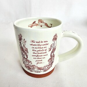May include: Cream ceramic mug with a brown base and handle. The mug features a mermaid illustration in brown ink, along with the text "Far out to sea where the water is as blue as the petals of the loveliest cornflower and as clear as purest glass..."