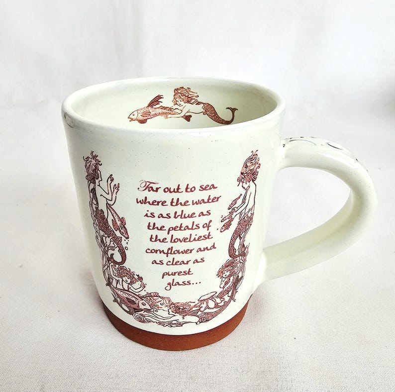 May include: Cream ceramic mug with a brown base and handle. The mug features a mermaid illustration in brown ink, along with the text "Far out to sea where the water is as blue as the petals of the loveliest cornflower and as clear as purest glass..."