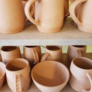 May include: A selection of terracotta mugs and bowls on shelves. The mugs feature large handles, some with spiral detailing. The unglazed pottery has a warm, earthy hue, giving a rustic or artisanal feel. The items are likely handmade.