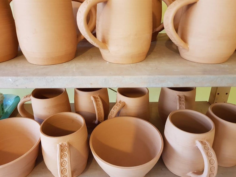 May include: A selection of terracotta mugs and bowls on shelves. The mugs feature large handles, some with spiral detailing. The unglazed pottery has a warm, earthy hue, giving a rustic or artisanal feel. The items are likely handmade.