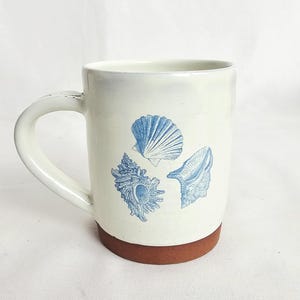 May include: Cream ceramic mug with a brown base and a white handle. The mug features three blue seashell illustrations. The mug is designed for hot drinks and is suitable for home or office use.