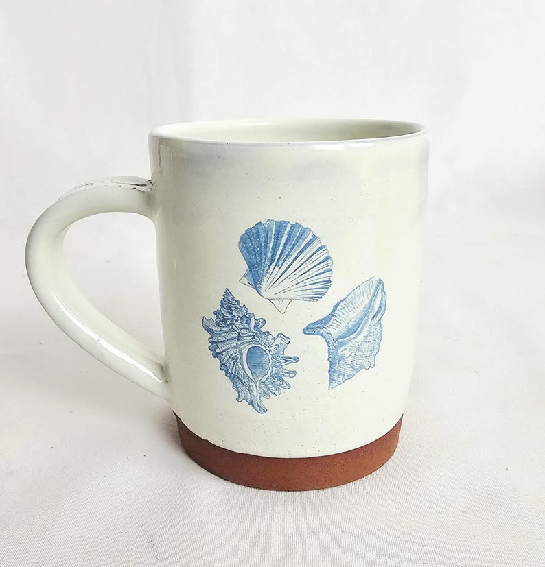 May include: Cream ceramic mug with a brown base and a white handle. The mug features three blue seashell illustrations. The mug is designed for hot drinks and is suitable for home or office use.