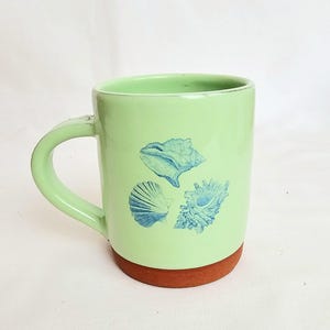 May include: A light green ceramic mug with a brown base and a curved handle. The mug features three blue seashell illustrations. The mug is designed for hot beverages and is suitable for home or office use.
