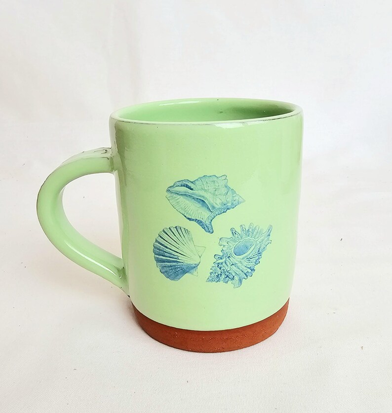 May include: A light green ceramic mug with a brown base and a curved handle. The mug features three blue seashell illustrations. The mug is designed for hot beverages and is suitable for home or office use.