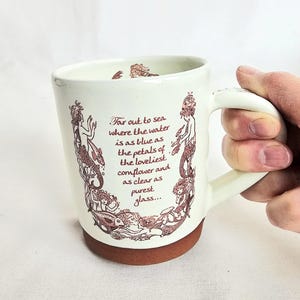 May include: Cream ceramic mug with a brown base and handle. The mug features a maroon illustration of mermaids and sea creatures, with the quote "Far out to sea where the water is as blue as the petals of the loveliest cornflower and as clear as purest glass..."