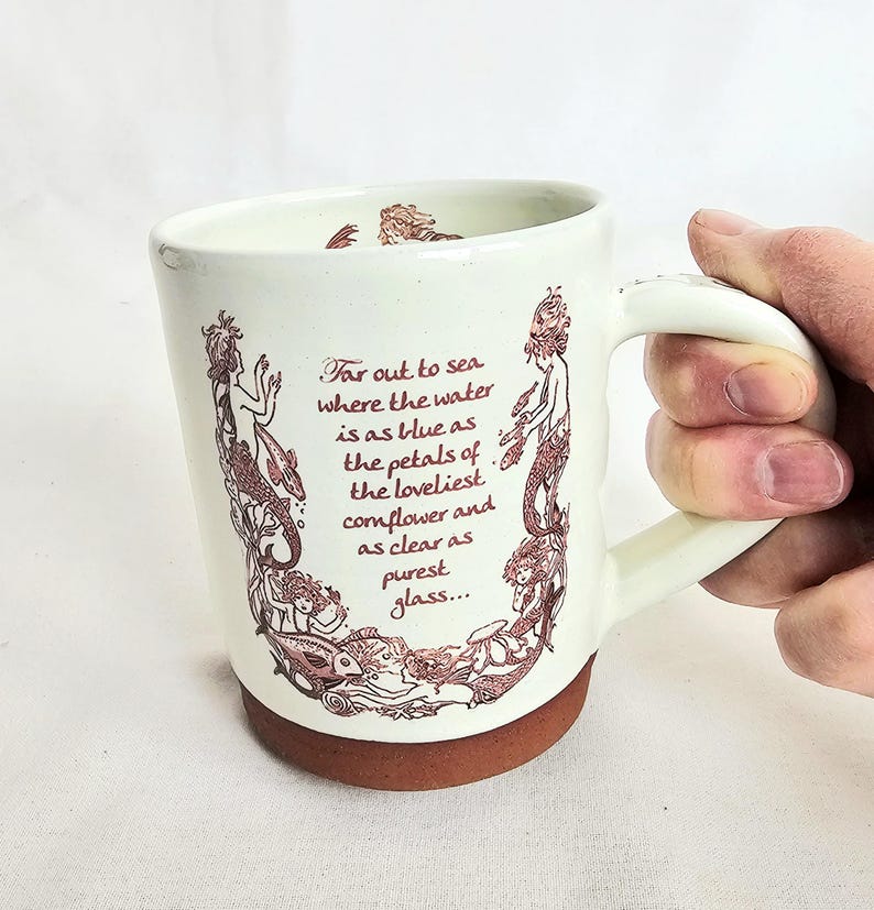May include: Cream ceramic mug with a brown base and handle. The mug features a maroon illustration of mermaids and sea creatures, with the quote "Far out to sea where the water is as blue as the petals of the loveliest cornflower and as clear as purest glass..."
