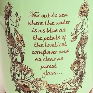 May include: A light green ceramic pot with a brown rim, featuring illustrations of mermaids and fish. The text reads, "Far out to sea where the water is as blue as the petals of the loveliest cornflower and as clear as purest glass..."