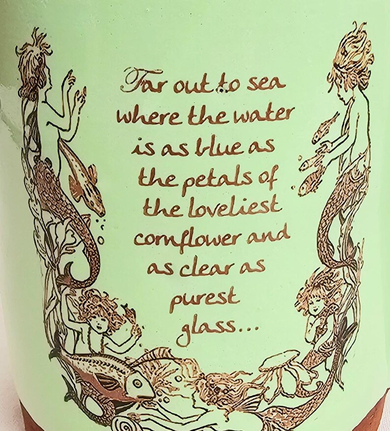 May include: A light green ceramic pot with a brown rim, featuring illustrations of mermaids and fish. The text reads, "Far out to sea where the water is as blue as the petals of the loveliest cornflower and as clear as purest glass..."