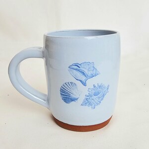 May include: A light blue ceramic mug with a brown base and a curved handle. The mug features three blue seashell illustrations. The mug is approximately 4 inches tall.