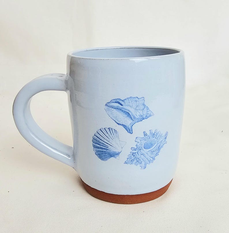 May include: A light blue ceramic mug with a brown base and a curved handle. The mug features three blue seashell illustrations. The mug is approximately 4 inches tall.