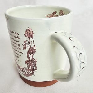 May include: Cream ceramic mug with a mermaid illustration in brown ink. The mug has a wide handle with a textured design and a brown base. The interior has a fish illustration. Text is printed on the side.