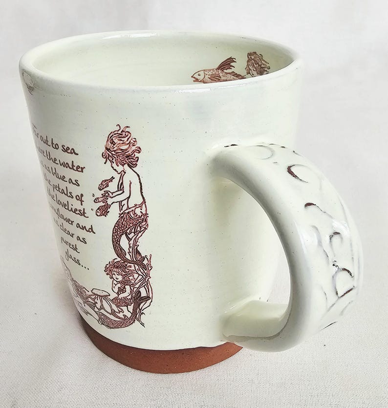 May include: Cream ceramic mug with a mermaid illustration in brown ink. The mug has a wide handle with a textured design and a brown base. The interior has a fish illustration. Text is printed on the side.