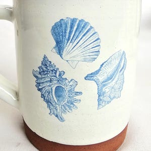 May include: Cream-coloured ceramic mug with a brown base and handle. The mug is decorated with three blue seashell designs: a scallop, a conch, and a clam. The mug has a rustic, handmade appearance.