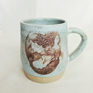 Könnte beinhalten: Ein Keramikbecher mit gesprenkelter hellblauer Glasur und einem naturfarbenen beigen Boden. Auf der Vorderseite befindet sich eine detaillierte braune Illustration einer Meerjungfrau mit langem Haar und geschwungenem Schwanz.