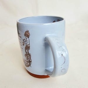 May include: A light blue ceramic mug with a brown base and a large handle. The mug features a mermaid illustration in brown ink with text. The mug is designed for hot beverages and is suitable for home or office use.