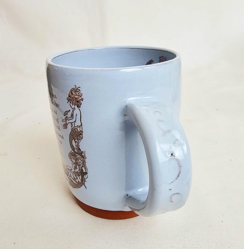 May include: A light blue ceramic mug with a brown base and a large handle. The mug features a mermaid illustration in brown ink with text. The mug is designed for hot beverages and is suitable for home or office use.