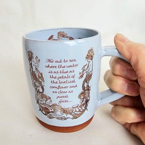 May include: A light blue ceramic mug with a brown base and handle. The mug features a design of mermaids and text in brown ink. The text reads: "Far out to sea where the water is as blue as the petals of the loveliest cornflower and as clear as purest glass..."