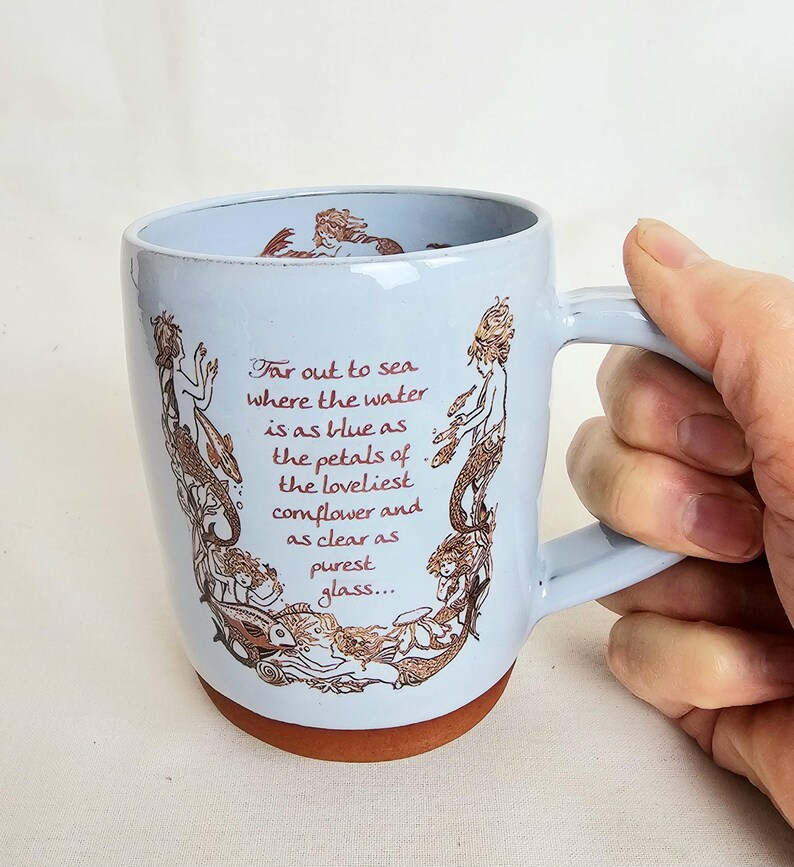May include: A light blue ceramic mug with a brown base and handle. The mug features a design of mermaids and text in brown ink. The text reads: "Far out to sea where the water is as blue as the petals of the loveliest cornflower and as clear as purest glass..."