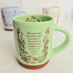 May include: A light green ceramic mug with a brown base and handle. The mug features a mermaid illustration and the text "Far out to sea where the water is as blue as the petals of the loveliest cornflower and as clear as purest glass..."