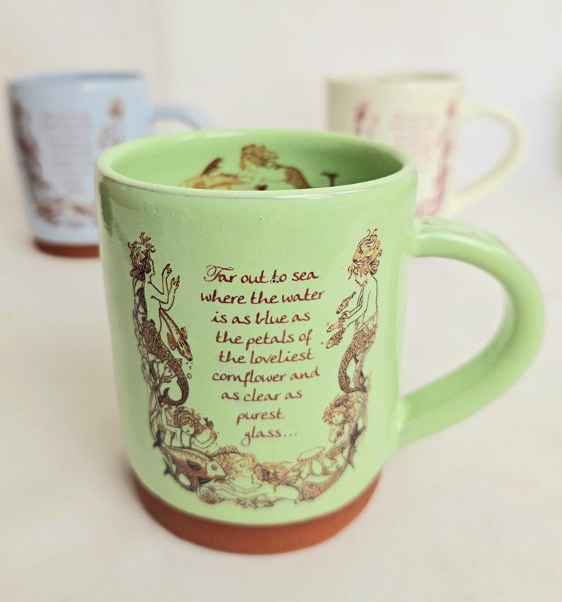 May include: A light green ceramic mug with a brown base and handle. The mug features a mermaid illustration and the text "Far out to sea where the water is as blue as the petals of the loveliest cornflower and as clear as purest glass..."
