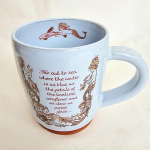 May include: A light blue ceramic mug with a brown base and handle. The mug features brown illustrations of mermaids and a fish, along with the text "Far out to sea where the water is as blue as the petals of the loveliest cornflower and as clear as purest glass..."