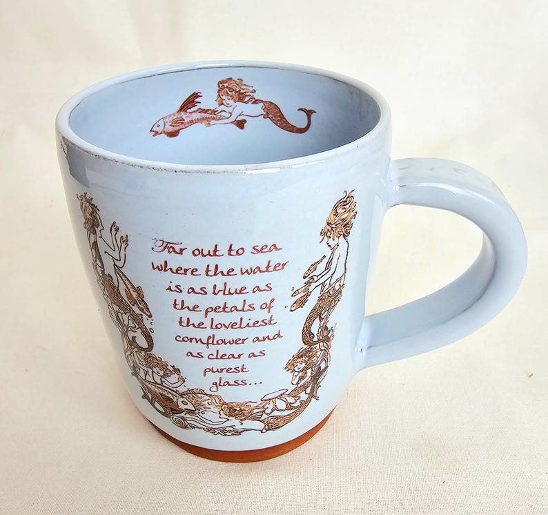 May include: A light blue ceramic mug with a brown base and handle. The mug features brown illustrations of mermaids and a fish, along with the text "Far out to sea where the water is as blue as the petals of the loveliest cornflower and as clear as purest glass..."