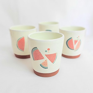Pode incluir: Quatro canecas de cerâmica com um esmalte amarelo claro e uma base marrom. Cada caneca apresenta um design diferente de fatia de melancia em tons de rosa, verde e azul. As canecas estão dispostas sobre uma superfície branca.