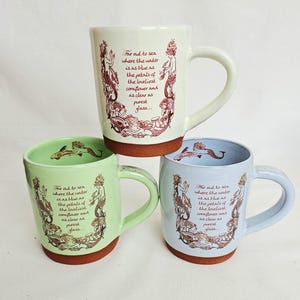 May include: Three ceramic mugs in cream, green, and blue, each with a brown base and handle. The mugs feature a mermaid illustration and the text "For out to sea where the water is as blue as the petals of the loveliest cornflower and as clear as purest glass..."