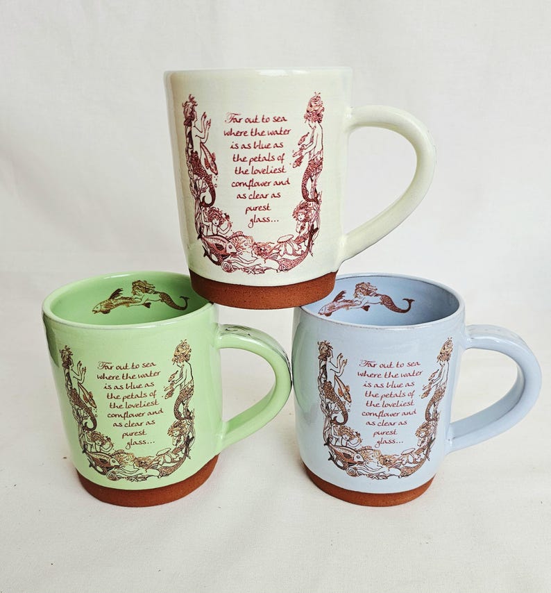 May include: Three ceramic mugs in cream, green, and blue, each with a brown base and handle. The mugs feature a mermaid illustration and the text "For out to sea where the water is as blue as the petals of the loveliest cornflower and as clear as purest glass..."