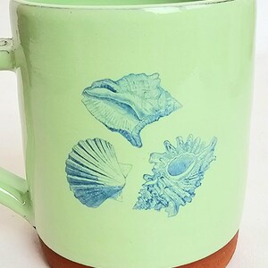 May include: A light green ceramic mug featuring three blue seashell illustrations. The mug has a brown base and a matching green handle. The design is suitable for a coastal or beach-themed kitchen.