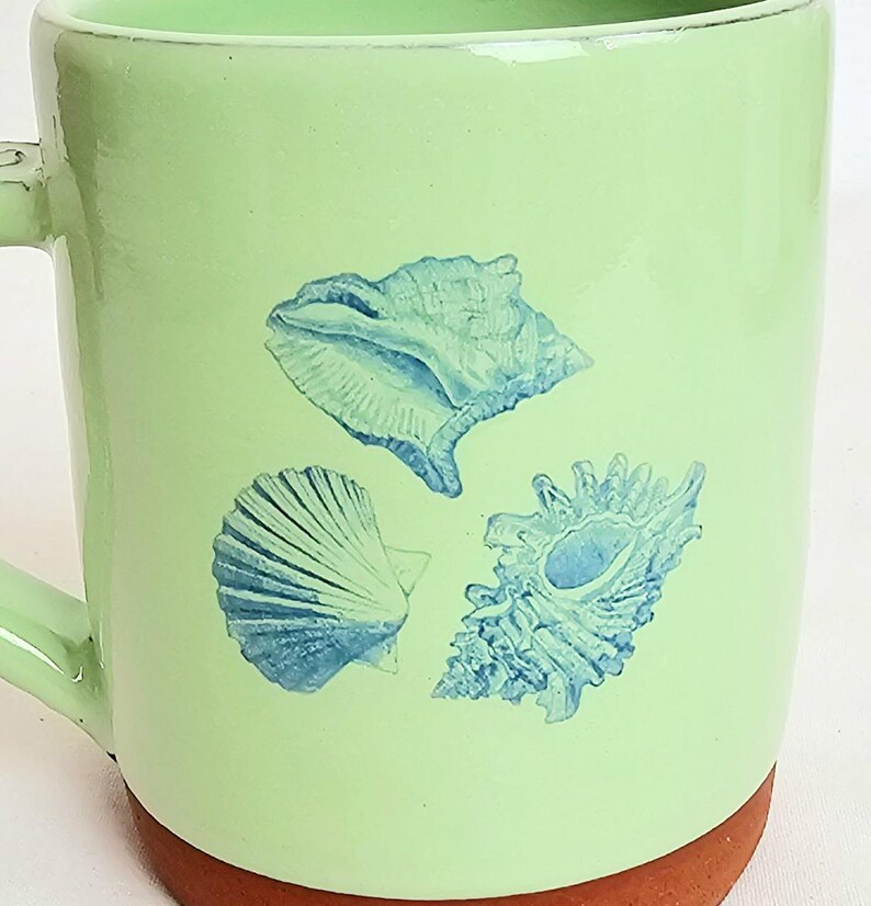 May include: A light green ceramic mug featuring three blue seashell illustrations. The mug has a brown base and a matching green handle. The design is suitable for a coastal or beach-themed kitchen.