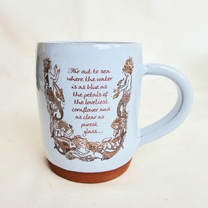 May include: A light blue ceramic mug with a brown base and handle. The mug features a decorative design of mermaids and the text "Far out to sea where the water is as blue as the petals of the loveliest cornflower and as clear as purest glass..."