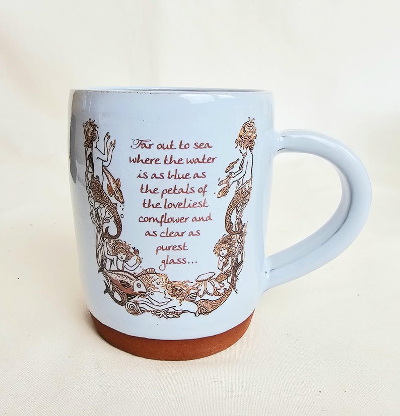 May include: A light blue ceramic mug with a brown base and handle. The mug features a decorative design of mermaids and the text "Far out to sea where the water is as blue as the petals of the loveliest cornflower and as clear as purest glass..."