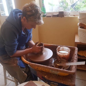 May include: A person using a pottery wheel to shape a clay pot. The wheel is brown and set in a yellow basin. The person wears a blue apron and hoodie. Buckets and pottery supplies are visible in the background.