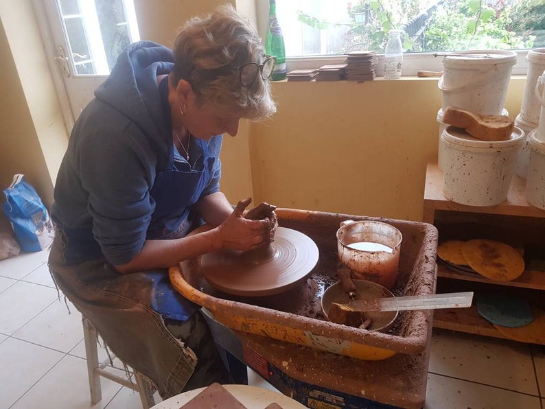 May include: A person using a pottery wheel to shape a clay pot. The wheel is brown and set in a yellow basin. The person wears a blue apron and hoodie. Buckets and pottery supplies are visible in the background.