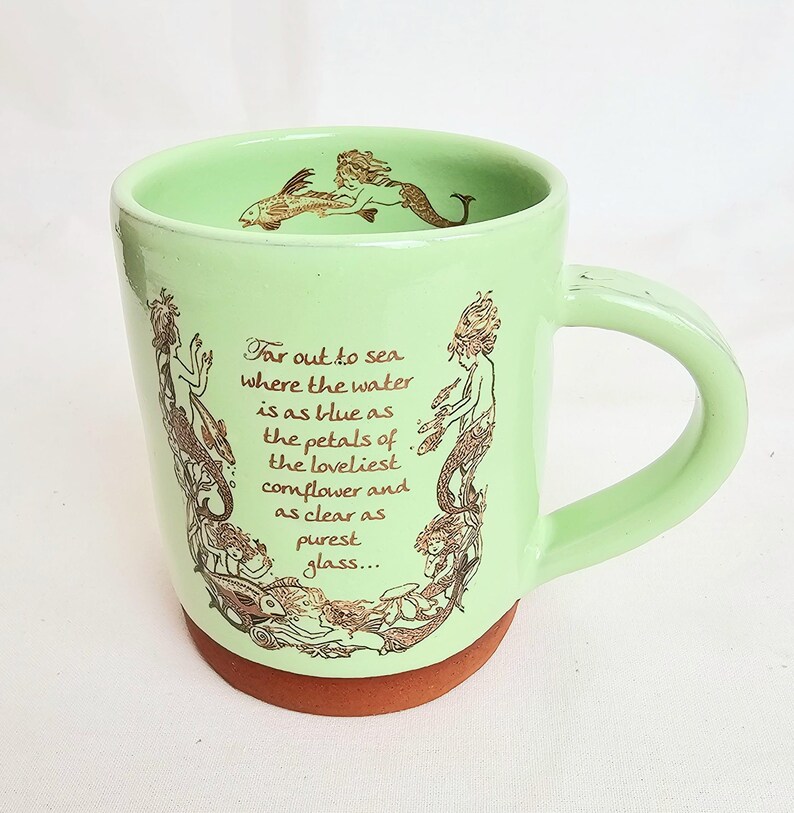 May include: A light green ceramic mug with a brown base and handle. The mug features a mermaid design in brown and the text "Far out to sea where the water is as blue as the petals of the loveliest cornflower and as clear as purest glass..."