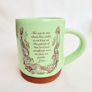 May include: A light green ceramic mug with a brown base and handle. The mug features a brown illustration of mermaids and fish, along with the text "Far out to sea where the water is as blue as the petals of the loveliest cornflower and as clear as purest glass..."