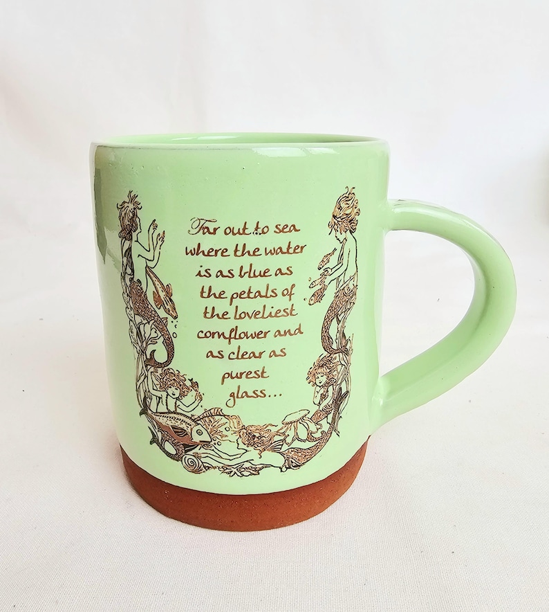 May include: A light green ceramic mug with a brown base and handle. The mug features a brown illustration of mermaids and fish, along with the text "Far out to sea where the water is as blue as the petals of the loveliest cornflower and as clear as purest glass..."