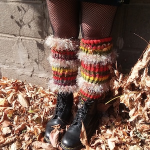 May include: A pair of black leather boots with laces. The boots are worn with colourful knitted leg warmers with a fluffy white trim. The leg warmers have red, orange, yellow, and green stripes. The boots and leg warmers are on a bed of fallen autumn leaves.