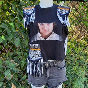 Puede incluir: Camiseta negra sin mangas con un gráfico de un hombre con sombrero. La camiseta está adornada con tela estampada y flecos. Se usa con pantalones cortos de mezclilla grises.