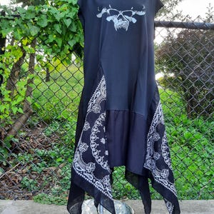May include: A black sleeveless dress featuring a white graphic print of a skull wearing sunglasses on the chest. The lower portion of the dress is constructed with black and white paisley patterned fabric panels that create an asymmetrical hemline.