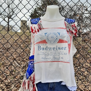 Puede incluir: Top sin mangas blanco con un diseño de etiqueta de cerveza Budweiser. La parte superior está adornada con detalles de tela roja, blanca y azul, y se combina con pantalones cortos de mezclilla. El dobladillo inferior de la parte superior tiene un ribete de encaje blanco.